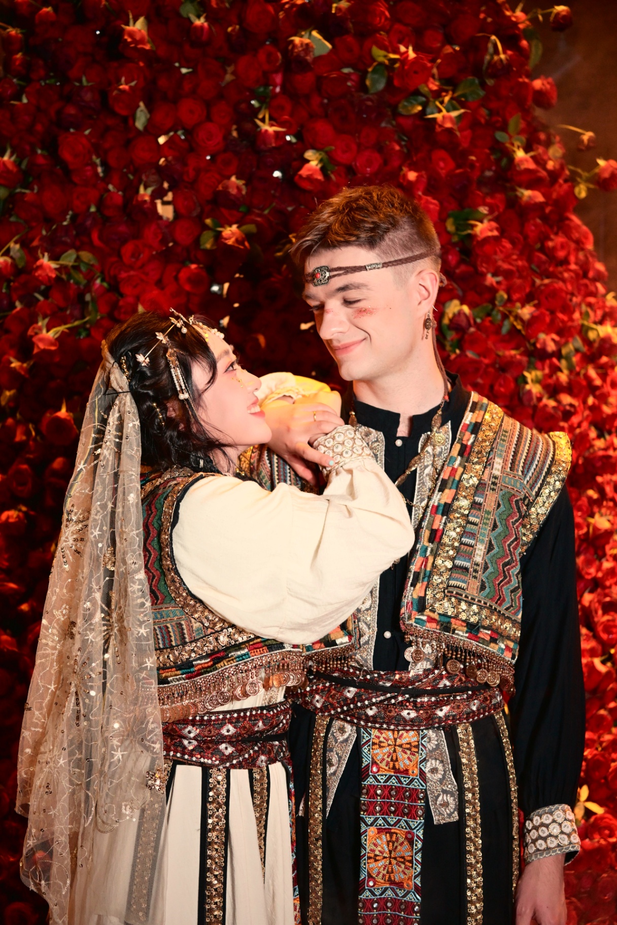 Oscar and Yan in traditional dress in front of a wall of red roses